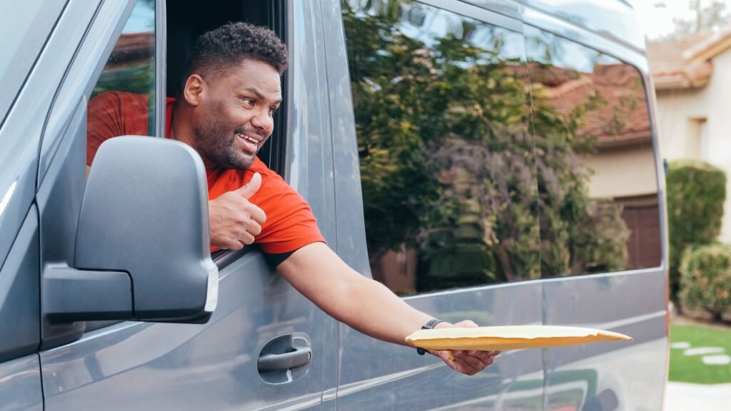 A delivery driver in a red shirt gives a thumbs up while handing a parcel from a van.